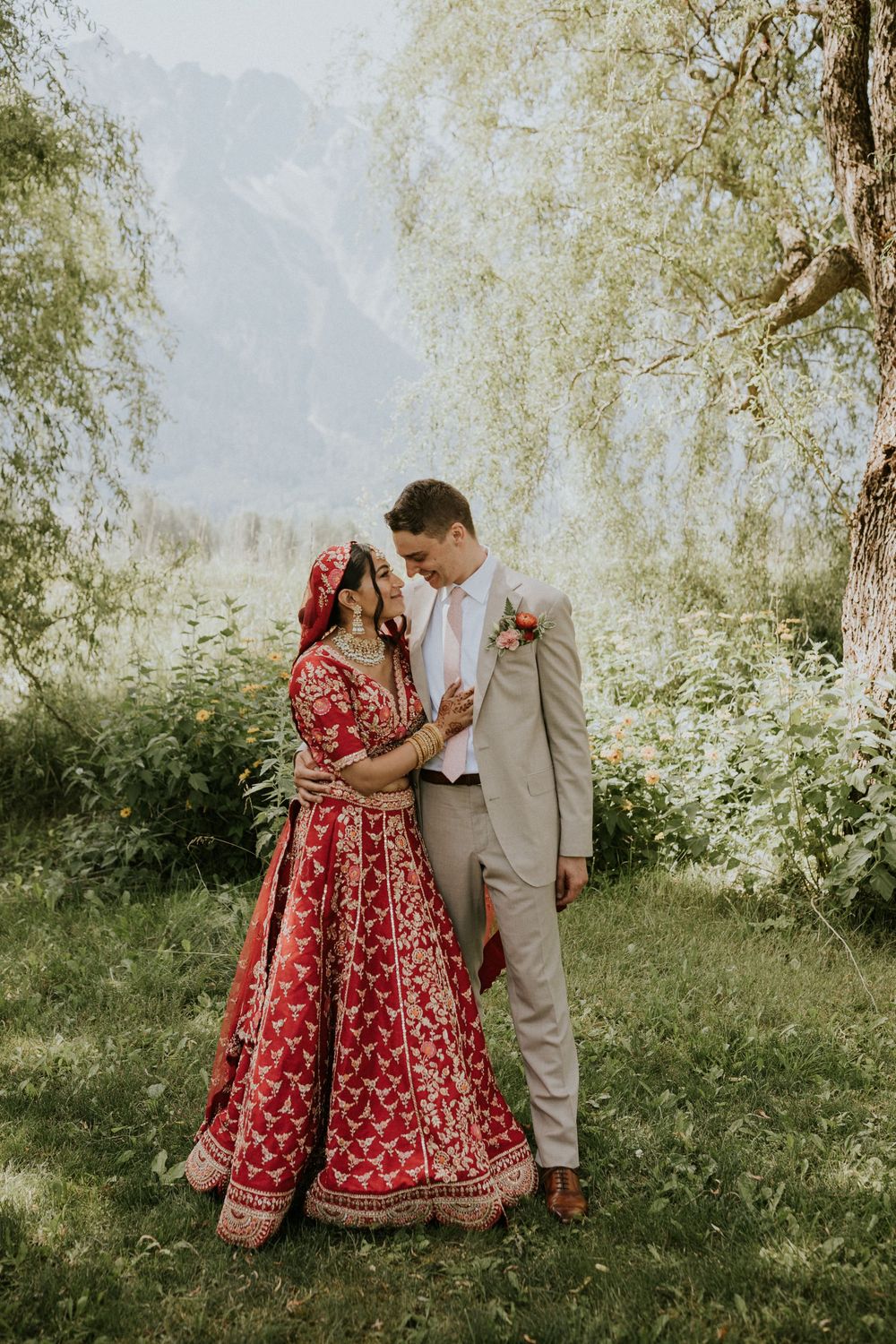 Wedding couple shares intimate moments in a garden setting with red traditional dress and beige suit.