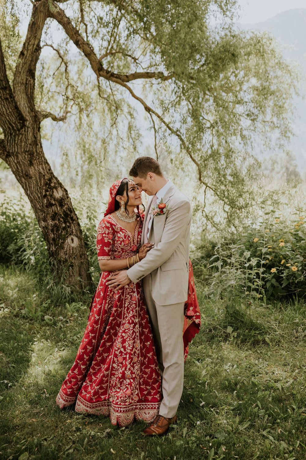 Romantic wedding portraits under willow trees with bride in red lehenga and groom in tan suit.