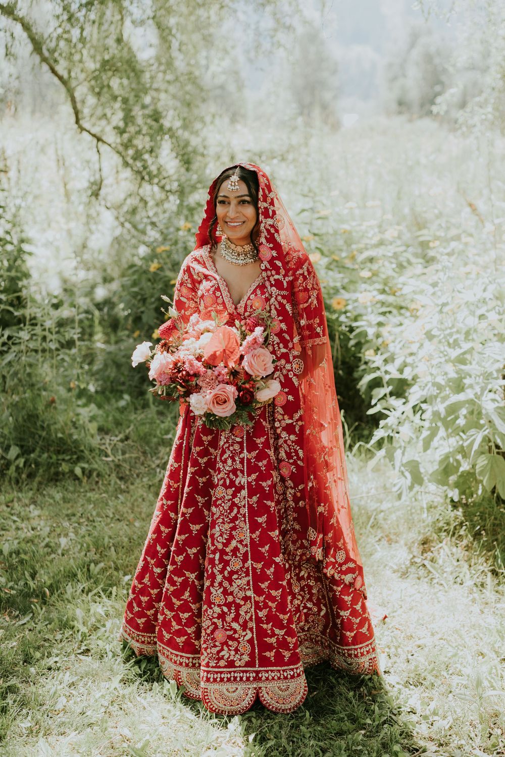 Traditional Indian bride in red embroidered lehenga holding pink floral bouquet against natural backdrop.