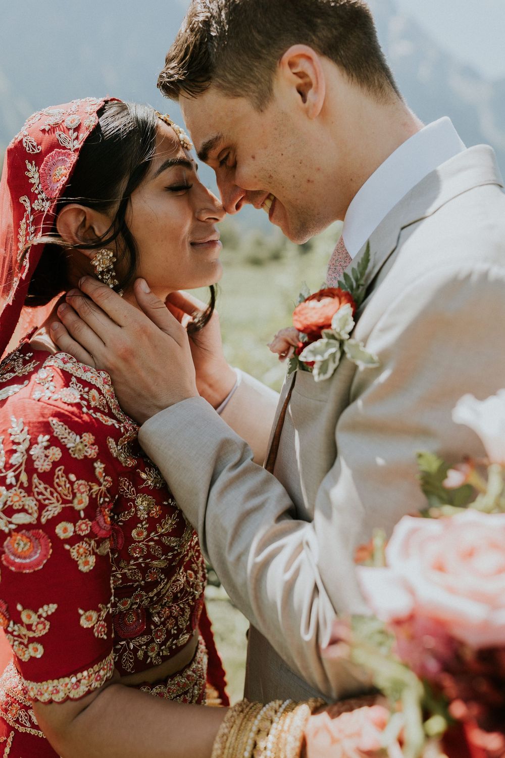A tender wedding moment showing a couple in traditional fusion attire with red and gold details against a mountain backdrop.