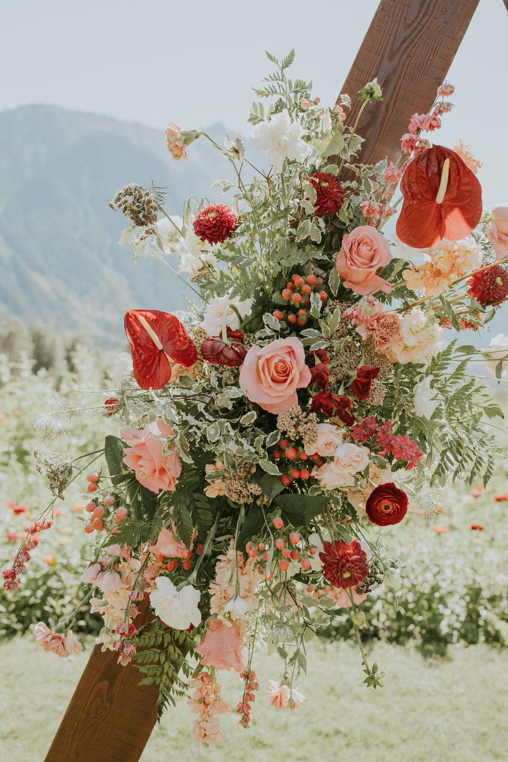 Rustic wooden arch decorated with lush coral and pink roses, greenery, and red dahlias against a mountain landscape.