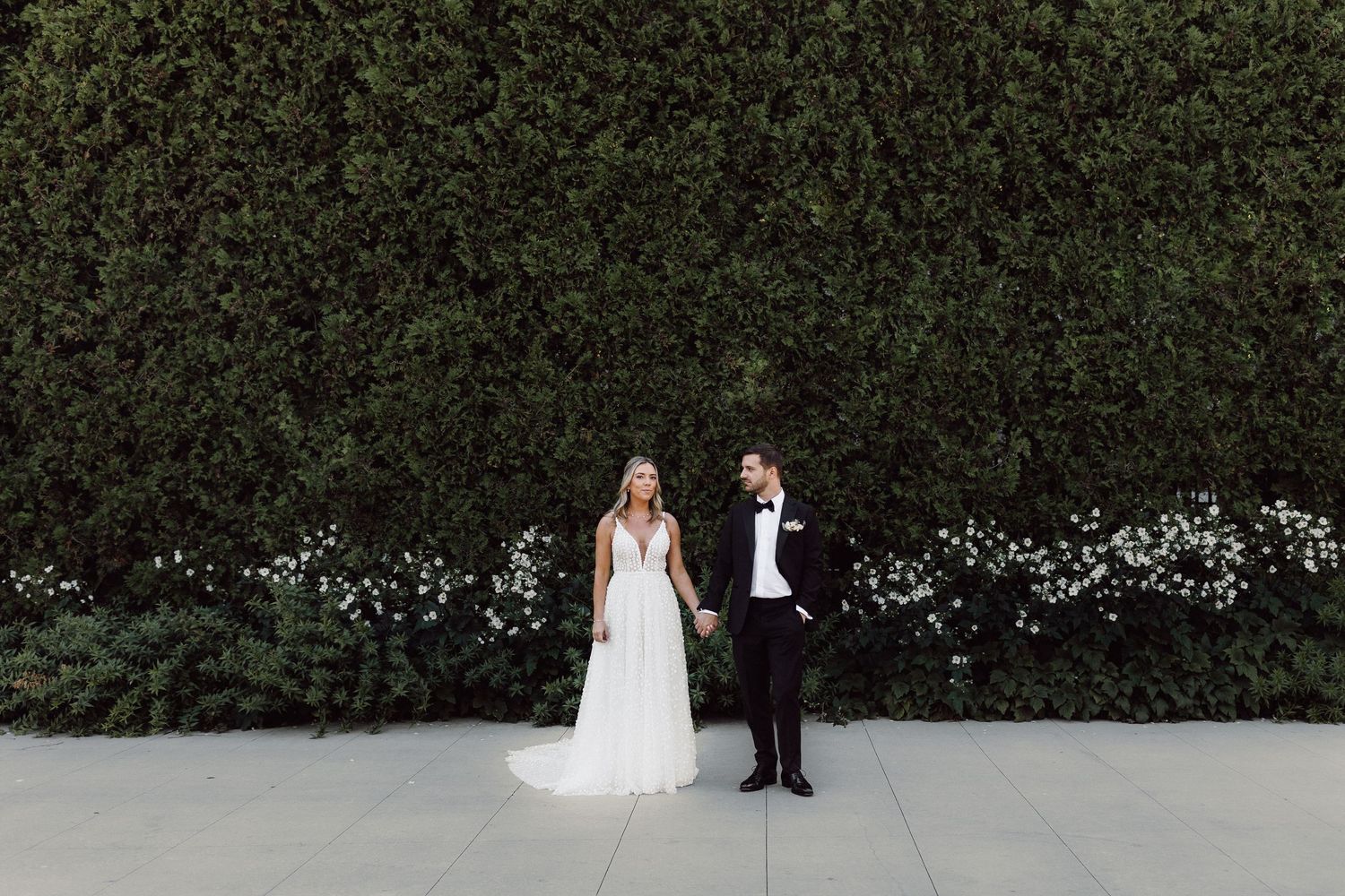 A couple in wedding attire walks through a lush garden path with tall hedges and white flowers.