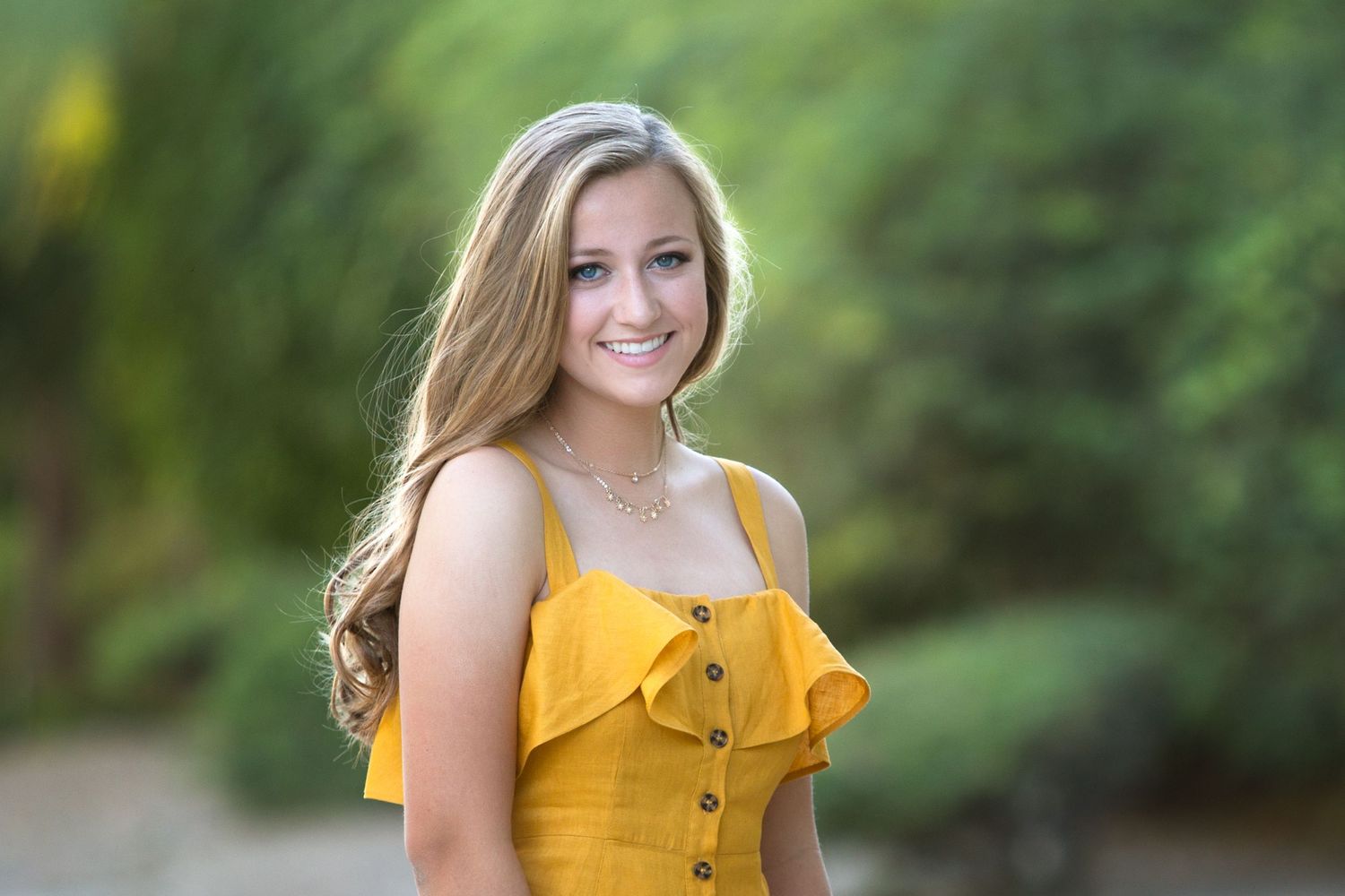 Woman in yellow summer top smiles while posing against blurred natural green background.