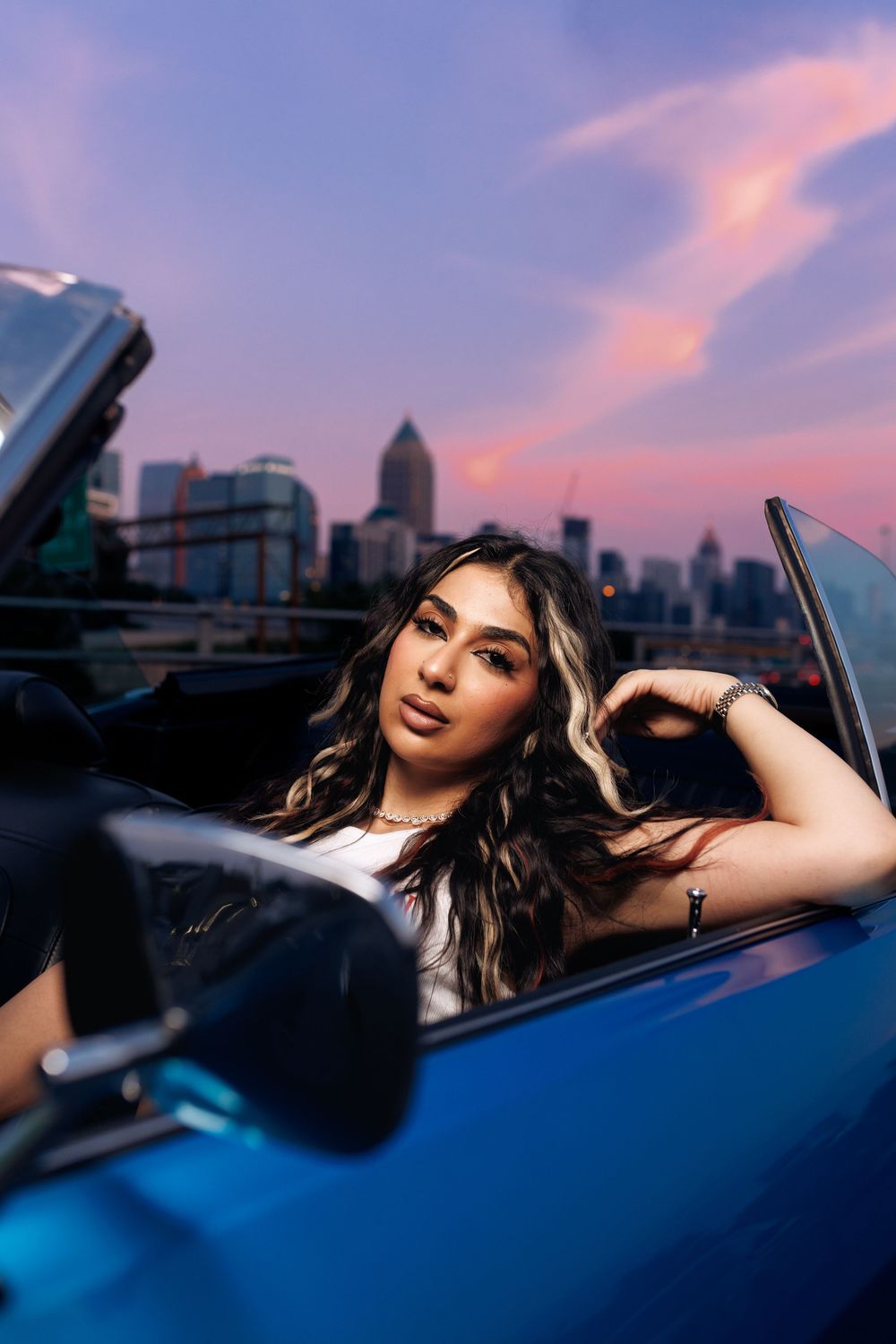 Stylish portrait in a blue convertible against a purple sunset city skyline.