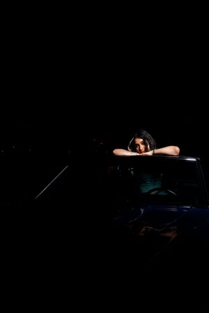 Dramatic low-light portrait leaning on blue car window against dark background.