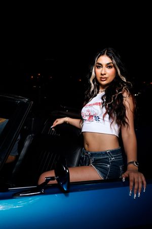 A model poses with a vintage car at night wearing denim shorts and a cropped white t-shirt.
