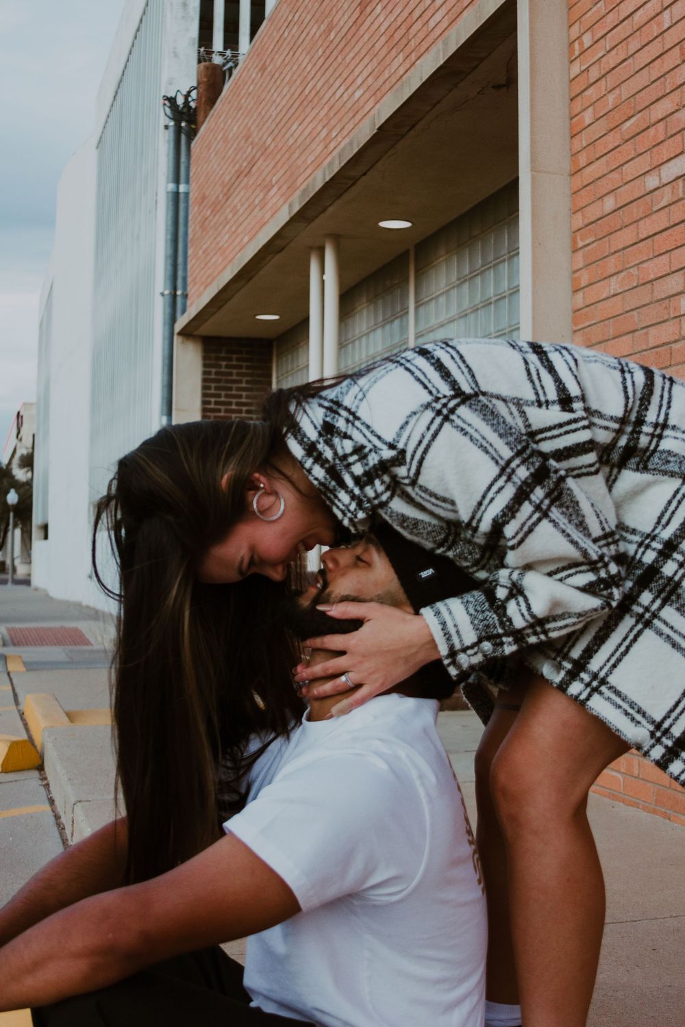 Woman in plaid jacket leaning down to kiss seated partner on urban sidewalk.