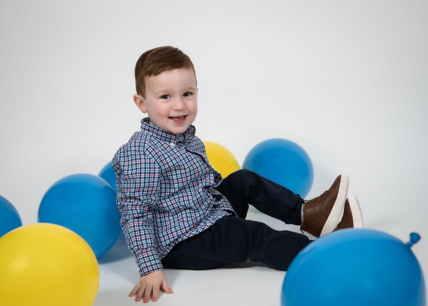 Toddler in plaid shirt and dark pants plays with blue and yellow balloons against white backdrop.