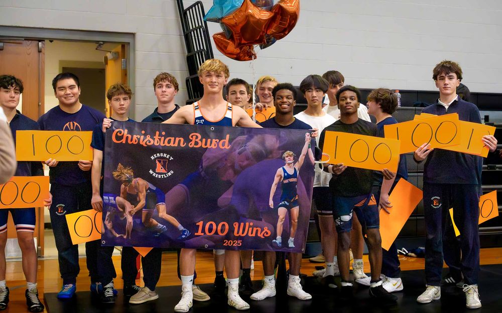 Hershey Wrestling team poses with Christian Burd and a “100 Wins” banner after his milestone victory.