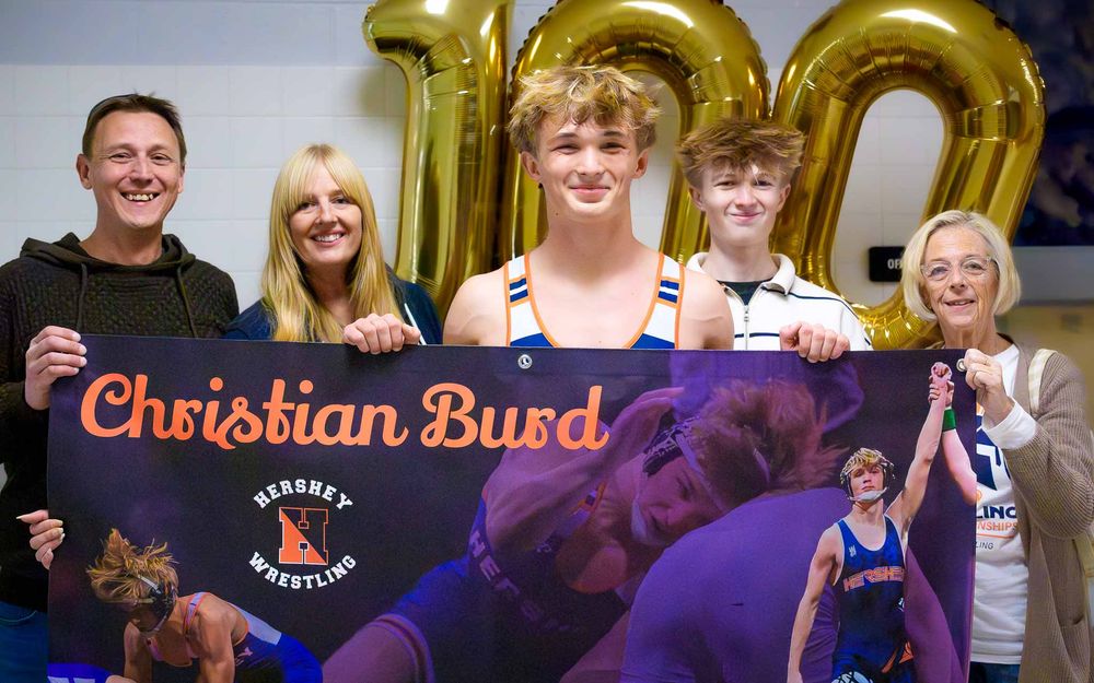 Christian Burd celebrates 100 career wrestling wins, holding a banner and surrounded by family and gold “100” balloons.