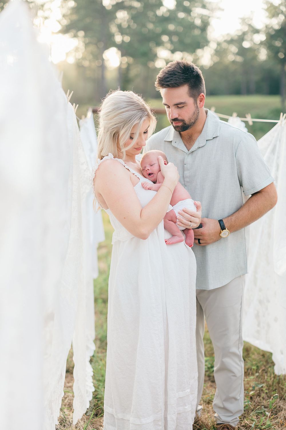 Couple holding newborn baby surrounded by hanging white sheets outdoors, captured by Mobile, AL Newborn Photographer.