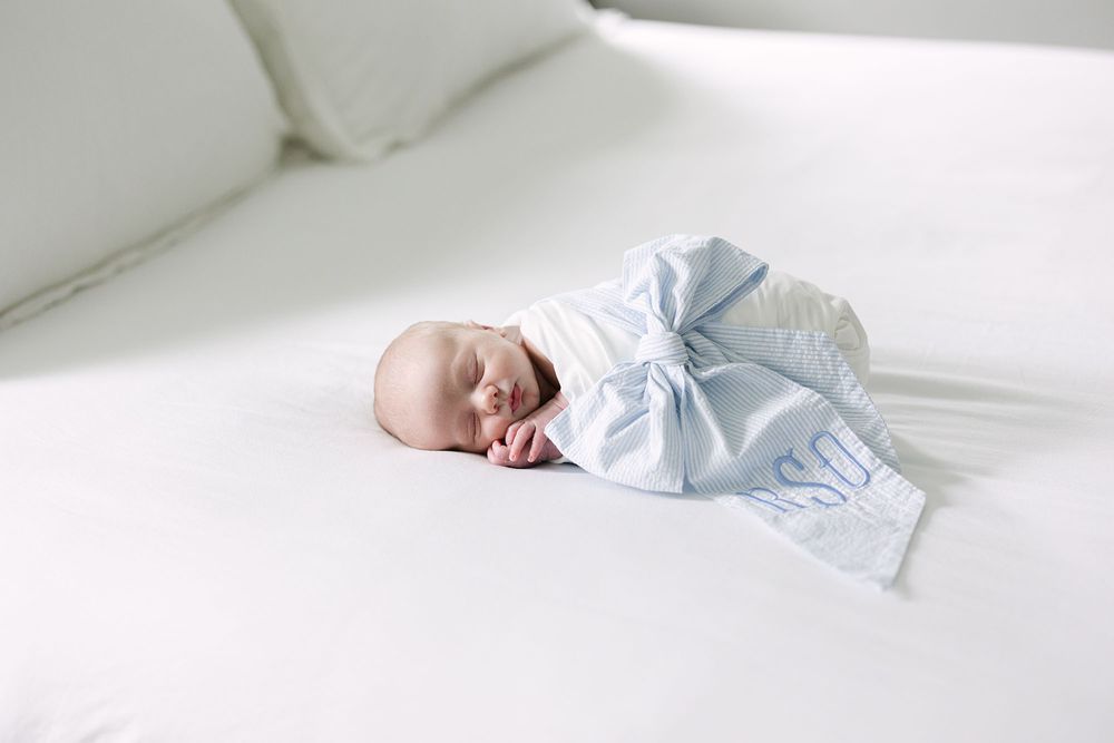 Baby swaddled in a white blanket with a blue bow sleeping on a white bed.
