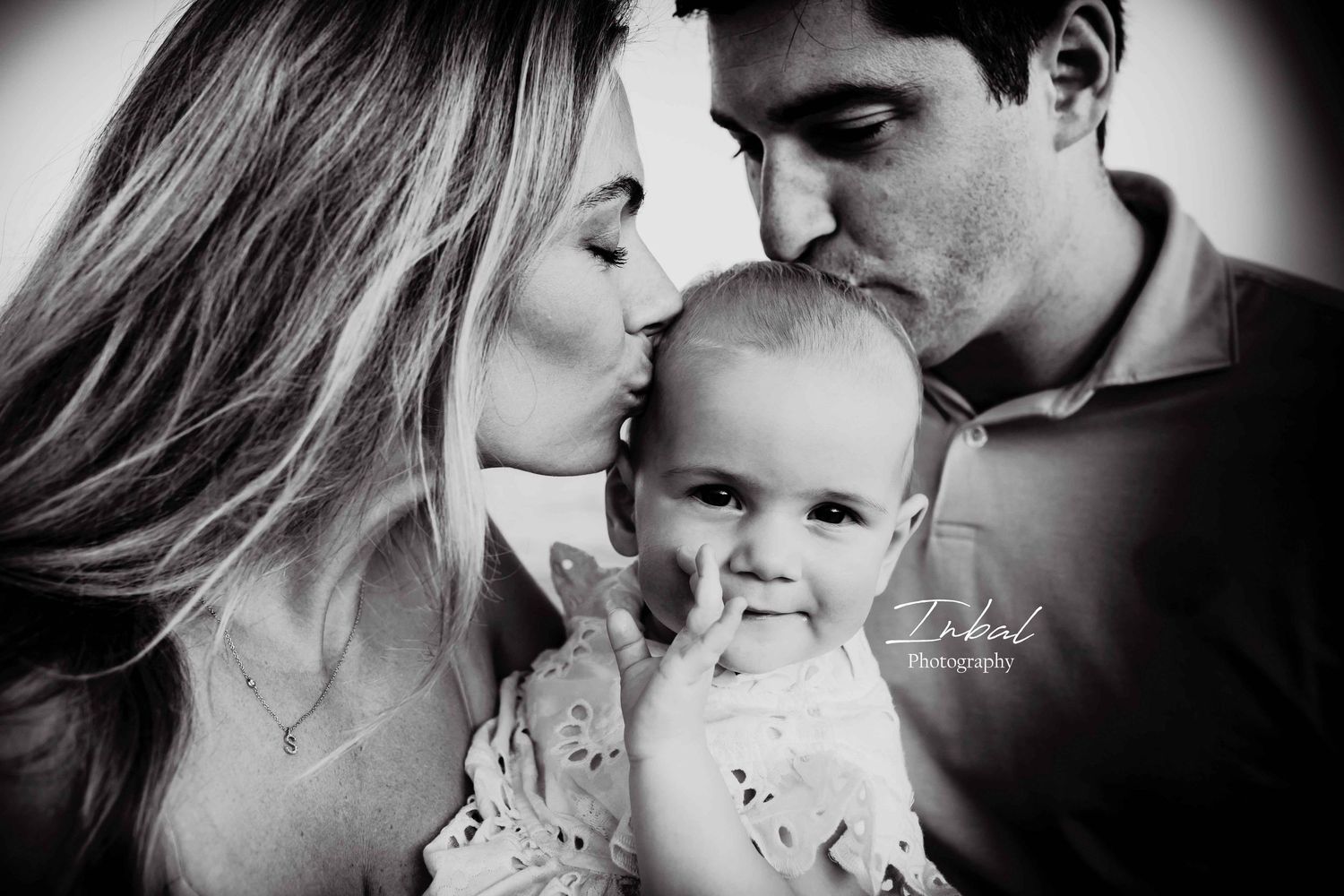Black-and-white photo of parents kiss their toddler girl head while she looks at the camera and smiling.