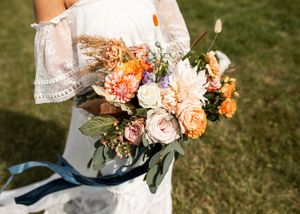 Bride holding a vibrant boho wedding bouquet with peach dahlias, roses, and dried grasses with blue ribbon.