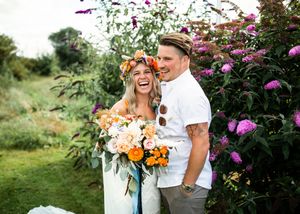 Laughing bride in floral crown and groom embracing before purple butterfly bush in colorful garden.