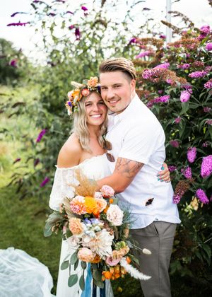 Smiling bride and groom posing with colorful boho bouquet and flower crown before purple blooms.
