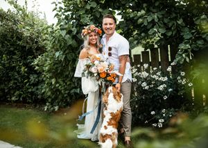Bride in floral crown and groom smiling with their spotted dog amid a lush green garden setting.