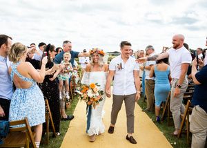 Bride with colorful floral crown and groom in white shirt walk through confetti-throwing guests at outdoor wedding.