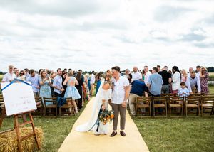 Newlywed couple walks down yellow aisle runner at outdoor farm wedding with hay bales and seated guests.