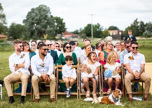 Wedding guests seated outdoors smiling with a dog sitting at their feet during ceremony.