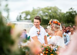 Groom in white shirt smiles at flower-crowned bride during outdoor wedding ceremony with seated guests behind them.