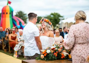Groom placing flower crown on bride during colorful outdoor wedding ceremony with bouncy castle.