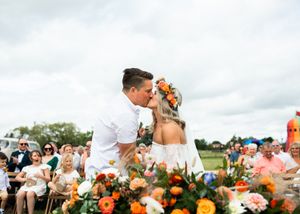 Bride and groom share first kiss at outdoor wedding ceremony surrounded by vibrant orange and white floral arrangements.