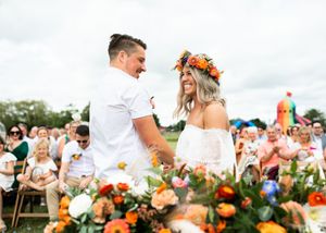 Groom and flower-crowned bride exchange vows at outdoor ceremony with colorful floral arrangements and seated wedding guests.