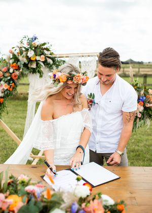 Bride signs wedding register wearing floral crown and off-shoulder dress while groom looks on at outdoor ceremony.