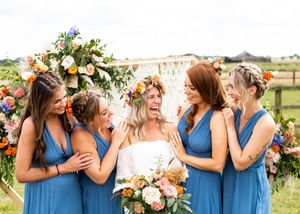 Bride in floral crown laughs joyfully with four bridesmaids in blue dresses at outdoor boho wedding.