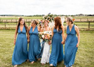 Bride holding colorful bouquet walks with four bridesmaids in blue maxi dresses at outdoor farm wedding.