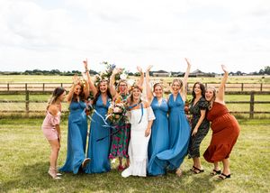 Bride celebrates joyfully with eight bridesmaids in blue and colorful dresses at outdoor farm wedding.