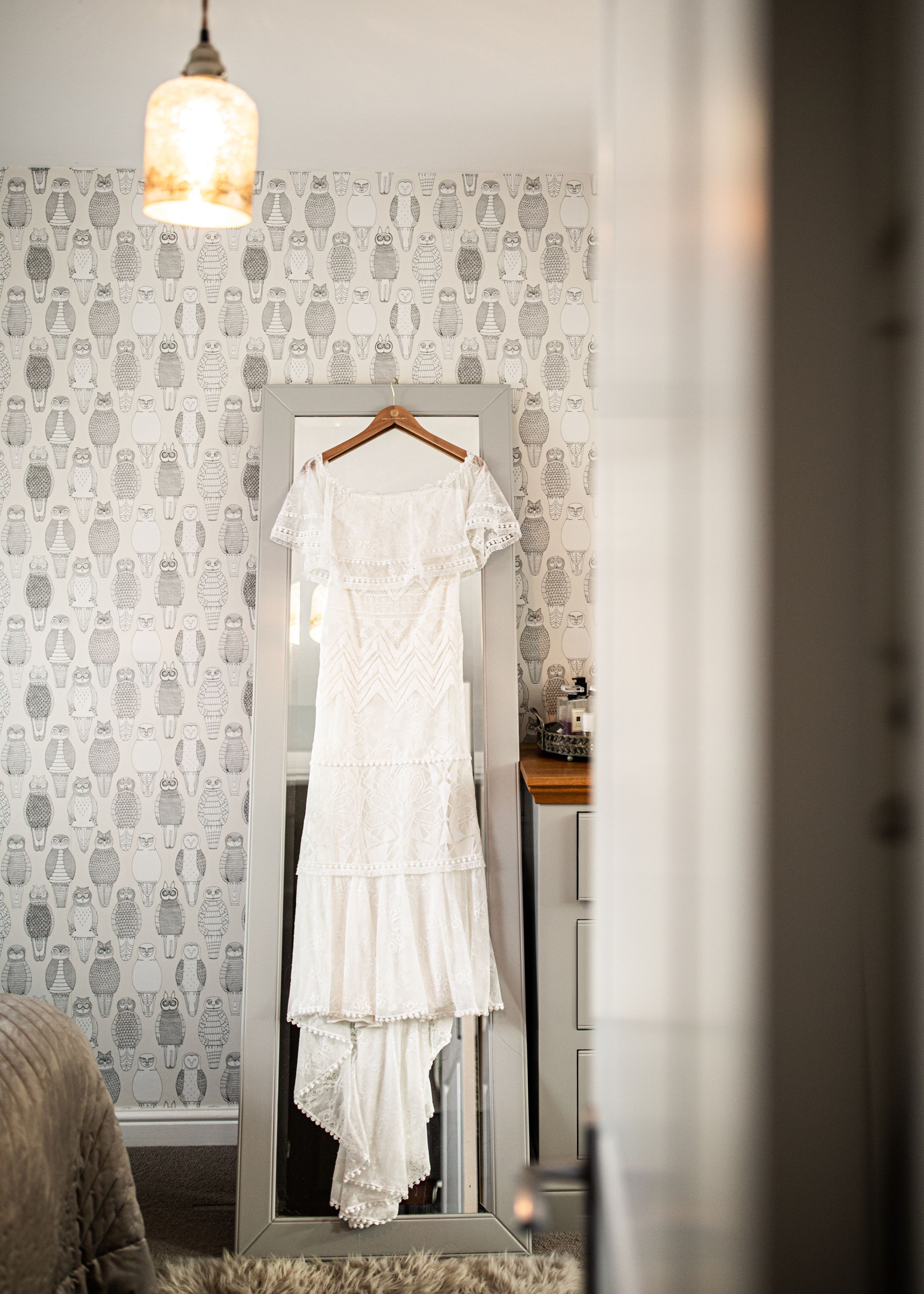 A white lace wedding dress hangs on a door in a room with decorative owl-patterned wallpaper.
