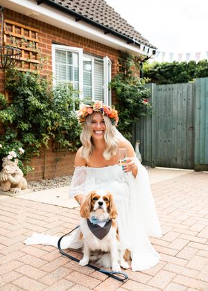 Blonde bride in floral crown and off-shoulder gown posing with Cavalier King Charles Spaniel outside.