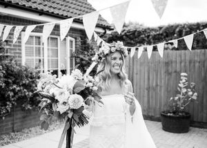 Smiling bride wearing floral crown holds bouquet at outdoor wedding with bunting decorations in black and white photo.