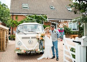 Bride and groom posing beside vintage VW camper van decorated with floral garlands outside venue.