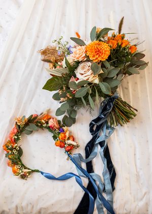 A bridal bouquet of orange dahlias and roses with blue ribbon beside a floral flower crown.