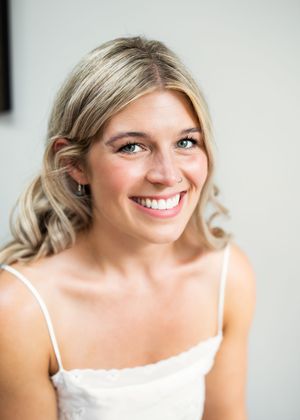 Smiling blonde bride with soft curled hair and natural makeup wearing a white spaghetti strap wedding dress.