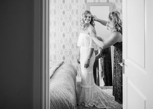 Black and white photo of bride in lace wedding gown getting hair styled by bridesmaid in decorated bedroom.