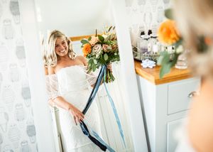 Blonde bride in strapless lace gown admiring herself in mirror while holding colorful orange floral bouquet.