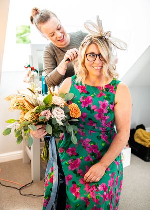 Woman in green floral dress holding bouquet getting fascinator styled by hairdresser in bright room.