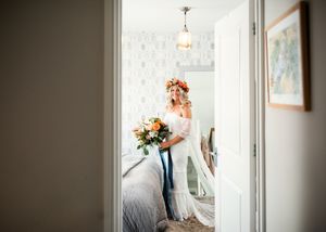 Bride in off-shoulder lace gown wearing floral crown holding bouquet viewed through bedroom doorway.
