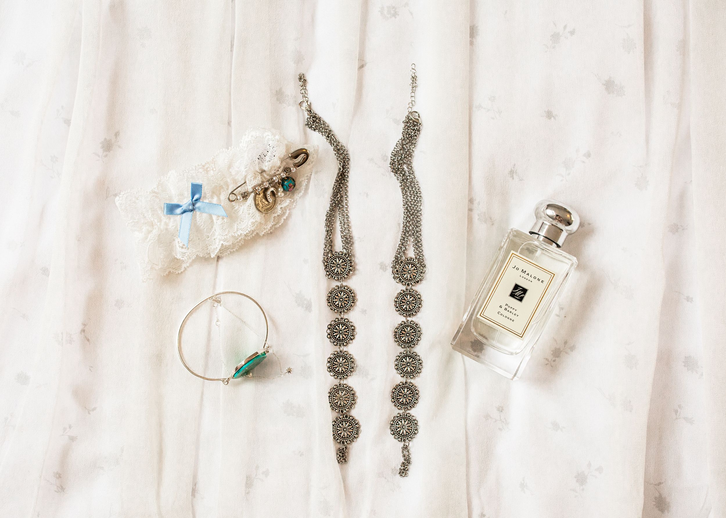 Flat lay of bridal accessories including jewelry, a garter, and Jo Malone perfume on white fabric.