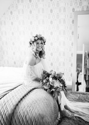 Black and white photo of smiling bride in floral crown sitting on bed holding large wedding bouquet.