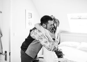 Black and white photo of emotional bride and suited groom sharing heartfelt hug in bright bedroom.