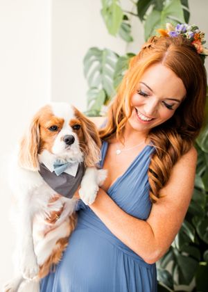 Smiling redhead bridesmaid in blue gown holding adorable Cavalier King Charles Spaniel in bow tie.