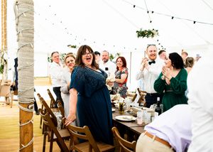 Wedding guests laughing and applauding inside a decorated marquee tent with fairy lights during a wedding reception.