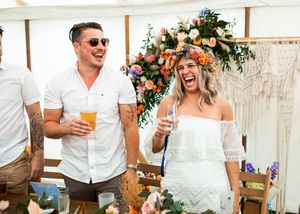 Laughing couple holding drinks at a boho wedding reception with floral backdrop.