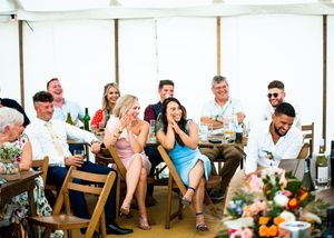 Wedding guests seated at long tables laughing during speeches inside a marquee tent.