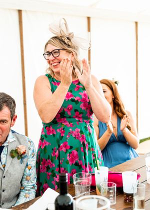 Woman in green floral dress and fascinator hat clapping joyfully at a wedding reception.